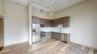 Unit 414 is a thoughtfully designed loft-style space that blends industrial character with contemporary finishes across a single open floor. Soaring ceilings with exposed structural elements and painted beams lend the space a distinctive industrial charm, while light-toned hardwood flooring flows seamlessly throughout, creating a warm and cohesive aesthetic.

The kitchen is a standout feature, offering an L-shaped layout equipped with a full suite of stainless steel appliances, including a French door refrigerator with ice maker, slide-in range, built-in microwave, and dishwasher. Rich, wood-grain cabinetry extends to the ceiling, maximizing storage, while white subway tile backsplash and light quartz countertops provide a clean, modern contrast.

The open living area is generously proportioned, bathed in natural light from a series of tall, multi-pane windows that frame urban views of the surrounding brick architecture. The airy volume of the space is enhanced by the dramatic ceiling height, giving the interior a bright and expansive feel.

The bathroom is well-appointed with a modern vanity featuring an integrated sink, framed mirror, and a walk-in glass shower. A stacked washer and dryer unit is conveniently integrated into the bathroom, adding practical in-unit laundry. Light tile flooring completes the clean, functional design of this well-rounded space.