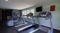 This single-floor fitness center is designed to support an effective and comfortable workout environment. The space features a dark rubberized flooring that provides both durability and safety underfoot, complemented by soft sage green and slate blue painted walls that create a calm, focused atmosphere. Recessed ceiling lighting keeps the room well-lit, while multiple windows along the far wall invite ample natural light and offer views of the surrounding greenery, adding a refreshing outdoor connection to the interior. The cardio equipment lineup includes multiple treadmills and elliptical cross-trainers, all positioned to face a wall-mounted flat-screen television, making it easy to stay entertained during workouts. Additional exercise equipment is visible along the walls, offering a range of training options. White baseboards and clean wall finishes give the space a tidy, well-maintained appearance. Overall, the fitness center strikes a balance between functionality and comfort, providing residents or users with a practical and pleasant space to meet their health and wellness goals.