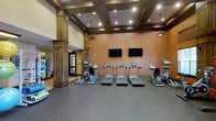 This fitness center is a thoughtfully designed workout space that combines functionality with refined architectural details. The room features soaring ceilings adorned with a striking coffered design framed in rich dark wood, complemented by recessed lighting and pendant fixtures that provide ample illumination throughout.

The workout floor is finished with durable rubber flooring in a neutral gray tone, ideal for supporting heavy equipment and high-traffic use. The warm terracotta-hued walls create an energizing atmosphere, while two wall-mounted flat-screen televisions are positioned for easy viewing from the cardio equipment area.

The cardio zone is well-stocked with a row of treadmills as the centerpiece, flanked by elliptical cross-trainers and stair-climbing machines, offering users a comprehensive range of aerobic workout options. A prominent decorative column anchors one corner of the room, adding an architectural element that echoes the upscale design aesthetic carried throughout the space.

An adjacent area, partially visible through a wide opening, suggests additional amenities nearby. The overall design balances a welcoming, club-like ambiance with the practical demands of a modern fitness facility, making it a well-rounded space suited for a variety of exercise routines.