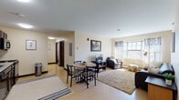 Located at 59 Broad St in Plattsburgh, New York, this single-floor unit offers a functional and comfortable layout designed to maximize livable space. The open living and dining area serves as the heart of the unit, where a round dining table with seating flows seamlessly into a carpeted living room furnished with a sofa, armchair, and television. Natural light streams through large windows dressed with sheer curtains, creating a bright and welcoming atmosphere throughout.

The kitchen is well-equipped with dark wood cabinetry, black appliances including a full-size refrigerator, dishwasher, electric range, and microwave, complemented by light-toned countertops and tile flooring. The space provides ample storage and prep area for everyday cooking needs.

Two private bedrooms each feature carpeted floors, a window that admits natural light, and individual dresser and desk combinations, offering both comfort and practicality. The bathroom includes a dual-sink vanity with a wide mirror and bright overhead lighting, alongside a separate water closet with tiled flooring, providing a practical and organized layout. Warm, neutral tones carry throughout the unit, lending a cohesive and understated aesthetic to the entire space.