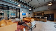This single-floor space showcases a refined industrial-modern aesthetic defined by exposed concrete ceilings, visible ductwork, and polished concrete floors. The open main lounge area is generously proportioned, anchored by a tufted cognac leather sofa, mixed seating arrangements, and a vibrant patterned area rug that adds warmth to the cool architectural palette. Pendant lighting and track fixtures provide layered illumination throughout.

Floor-to-ceiling black-framed glass windows and doors line one wall, flooding the interior with natural light and offering direct views of mature trees and an outdoor terrace with bar-height seating. The same black steel framing is used for interior glass partitions and corridor walls, creating a cohesive, loft-like visual language.

A dedicated kitchenette features warm wood cabinetry, open shelving, a white countertop, built-in microwave, and a full-size stainless refrigerator. Adjacent bar-height seating connects the kitchen area to the lounge. A separate conference room offers a dark metal table, seating for six, and a wall-mounted display screen. The corridor leading to private spaces is accented with large potted trees, adding a lush, biophilic element to the overall environment.