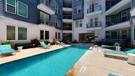 This modern residential unit is part of a multi-story building with a striking exterior characterized by dark blue siding and white trim, featuring private balconies. Residents enjoy access to a central courtyard with a sparkling outdoor pool, sun-drenched pool deck, and lounge seating — perfect for relaxing outdoors.

Inside, the open-concept layout flows naturally from a well-designed kitchen into the main living area. The kitchen is outfitted with rich dark wood cabinetry, white quartz countertops, a white subway tile backsplash, and a full suite of stainless steel appliances including a range, microwave, and dishwasher. Warm hardwood-style flooring runs throughout the main living areas, complemented by pendant and track lighting that adds a contemporary touch. A kitchen island provides additional prep and dining space.

The bedroom offers plush wall-to-wall carpeting, neutral gray walls, and large windows with blinds that invite natural light and frame outdoor views. A ceiling fan adds comfort to the space.

The bathroom features dark wood vanity cabinetry, a white countertop with an undermount sink, and hardwood-style flooring that carries over from the main living areas, maintaining a cohesive aesthetic throughout. The overall ambiance is clean, modern, and functional.