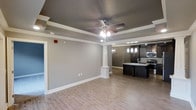 This well-appointed single-floor space showcases a thoughtfully designed open-concept layout that seamlessly connects the living area to the kitchen. The living space is defined by an elegant tray ceiling with a decorative border, complemented by a ceiling fan with integrated lighting. Light-toned hardwood flooring runs throughout, creating a warm and cohesive aesthetic. A decorative column with a classic pedestal base serves as a refined architectural detail, subtly delineating the living and kitchen areas without interrupting the open flow.

The kitchen is a standout feature, boasting rich dark cabinetry that provides a striking contrast against the neutral gray walls. A kitchen island with a sink anchors the space, while stylish pendant lights hang overhead to illuminate the workspace. A tile backsplash adds texture and visual interest, and stainless steel appliances complete the modern, functional setup. A globe chandelier in the transitional area between rooms adds an additional layer of ambient lighting and design character.

The neutral gray wall palette throughout creates a clean, contemporary backdrop that enhances the architectural details. The combination of tray ceilings, decorative columns, and carefully chosen fixtures gives this space a polished and inviting character suited for both everyday living and entertaining.