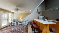 This single-floor space is designed as a multifunctional common area with a warm, lodge-inspired character. The lounge section features a pair of cushioned striped armchairs arranged around a small side table with a lamp, creating an inviting seating nook. Adjacent to the lounge is a built-in bar counter with warm wood cabinetry, a white laminate top, and red tufted bar stools. The bar area is equipped with a refrigerator and microwave, along with a coffee and beverage station. A mounted elk antler trophy adds a distinctive rustic accent above the bar. The space transitions into a game area with a table and chairs positioned beneath soaring ceilings with dual ceiling fans. French doors with glass panes connect to additional rooms, allowing natural light to flow through. A large white painted brick fireplace serves as a focal point along one wall, flanked by windows that overlook a landscaped exterior. Bulletin boards and signage near the fireplace suggest a community-oriented use. The flooring combines patterned carpet with tile, and the textured ceiling runs throughout, contributing to the cohesive, casual ambiance of the space.