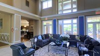Located at 400 Stone Lion Dr in Durham, NC, this single-floor community clubhouse offers an impressive and welcoming environment defined by exceptional architectural detail and thoughtful design. The main lounge area commands immediate attention with its soaring double-height ceilings and an expansive wall of windows that floods the space with natural light, offering views of the lush outdoor surroundings. Tall transom windows positioned above the main glazing further amplify the sense of openness and grandeur. The lounge is furnished with plush sectional sofas in deep blue-gray tones, complemented by accent chairs in rich navy leather, a glass-top coffee table, and floral arrangements that add a fresh, refined touch. A patterned area rug anchors the seating area, tying together the elegant aesthetic. A bold royal blue accent wall provides a striking architectural focal point, adding a vibrant contrast to the warm neutral tones of the surrounding walls. Adjacent to the lounge, a well-appointed dining and kitchen area is visible, featuring dark wood furnishings, pendant lighting, and a layout suited for gatherings and social events. Throughout the space, the combination of warm finishes, abundant daylight, and carefully curated furnishings creates an inviting and polished ambiance.