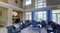 Located at 400 Stone Lion Dr in Durham, NC, this single-floor community clubhouse offers an impressive and welcoming environment defined by exceptional architectural detail and thoughtful design. The main lounge area commands immediate attention with its soaring double-height ceilings and an expansive wall of windows that floods the space with natural light, offering views of the lush outdoor surroundings. Tall transom windows positioned above the main glazing further amplify the sense of openness and grandeur. The lounge is furnished with plush sectional sofas in deep blue-gray tones, complemented by accent chairs in rich navy leather, a glass-top coffee table, and floral arrangements that add a fresh, refined touch. A patterned area rug anchors the seating area, tying together the elegant aesthetic. A bold royal blue accent wall provides a striking architectural focal point, adding a vibrant contrast to the warm neutral tones of the surrounding walls. Adjacent to the lounge, a well-appointed dining and kitchen area is visible, featuring dark wood furnishings, pendant lighting, and a layout suited for gatherings and social events. Throughout the space, the combination of warm finishes, abundant daylight, and carefully curated furnishings creates an inviting and polished ambiance.
