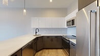 Located at 125 Warren St in Boston, MA, this thoughtfully designed single-floor residence combines contemporary finishes with functional living spaces. The open-concept kitchen serves as a central highlight, showcasing high-gloss white upper cabinetry, contrasting dark lower cabinets, a classic white subway tile backsplash, and bright quartz countertops. A generous peninsula with pendant lighting above creates an ideal space for dining or entertaining, while stainless steel appliances, including a range, microwave, and refrigerator, complete the modern aesthetic. Light-toned hardwood flooring flows seamlessly throughout the living areas, enhancing the sense of openness and warmth. The living and dining areas benefit from recessed lighting and a neutral color palette, creating an inviting, airy atmosphere. Multiple windows across the bedrooms invite an abundance of natural light and offer views of the surrounding Boston neighborhood, with the city's vibrant architecture visible from several vantage points. The bathroom is finished with large-format tile flooring, a white vanity with matte black hardware, and a tub-shower combination enclosed by sleek black-framed glass sliding doors, set against marble-look tile surround. The overall design of the space reflects a clean, contemporary aesthetic well-suited for urban living in the heart of Boston.