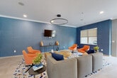 This single-floor community amenity space is thoughtfully designed to offer both relaxation and entertainment. The lounge area makes a bold visual statement with deep blue textured accent walls, crown molding, and light large-format tile flooring. A circular pendant light fixture anchors the seating arrangement, which features a generously sized sectional sofa in warm gray, complemented by vibrant orange swivel accent chairs. A vivid terrazzo-style area rug in blue, orange, and cream ties the color palette together, while a wall-mounted flat-screen TV and a sleek console table complete the media-ready setup. Abstract artwork in coordinating tones adds a modern, curated feel to the space. The adjacent games room shifts to a more casual, open atmosphere, highlighted by a full-size pool table, a shuffleboard table, and a wall-mounted TV set against a warm wood slat accent wall. A ceiling fan overhead and abundant recessed lighting keep the space comfortable and well-lit. Most notably, the games room features retractable glass garage doors that open fully to an outdoor pool and patio area, flooding the interior with natural light and seamlessly blending indoor and outdoor living. Together, these areas create an engaging, social environment well-suited for both leisure and community gatherings.