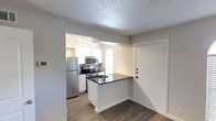 This single-story space showcases a well-designed open-concept kitchen and entry area. The kitchen is outfitted with stainless steel appliances, including a full-size refrigerator, electric range, and over-the-range microwave, all set against crisp white cabinetry that provides ample storage. A peninsula with a dark countertop serves as both a workspace and a natural divider between the kitchen and the adjacent living area, adding functionality to the layout.

The flooring throughout features dark wood-look planks, providing a modern contrast to the light-colored walls and white cabinetry. Track lighting in the kitchen ensures the workspace is well-illuminated, while an arched window above the sink allows natural light to filter in, brightening the cooking area. The textured popcorn ceiling is consistent throughout the visible areas.

The entry area, positioned adjacent to the kitchen, features a white front door and a neutral color palette that contributes to the overall clean and cohesive aesthetic of the space. The layout is compact yet efficient, making practical use of the available area.