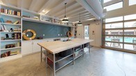 This expansive single-floor amenity space combines industrial architecture with warm, eclectic design to create a compelling communal environment. Soaring exposed beam ceilings stretch high overhead, amplifying the sense of openness throughout. A dramatic wall of floor-to-ceiling glass garage-style doors floods the lounge area with natural light while offering unobstructed views of an outdoor pool and surrounding grounds.

The main lounge features a curated seating arrangement — including a cream sofa, cognac leather armchairs, and plush emerald wingback chairs — anchored by a vibrant Persian-style rug and complemented by a wall-mounted television and open industrial bookshelves. Globe pendant lights are suspended in clusters from the ceiling, adding a warm, ambient glow.

Adjacent to the lounge, a generously sized billiards table anchors a games area beneath the same soaring ceiling, with bar-height seating, large-scale artwork, and a mezzanine-level overlook above a light wood staircase. Polished concrete floors run throughout, lending a sleek, cohesive industrial character to the space.

The kitchen area offers a large butcher block island with a stainless steel frame, teal subway tile backsplash, open shelving, and pendant lighting — a practical and stylish hub for communal gatherings. Exposed ductwork and structural beams throughout reinforce the space's modern industrial aesthetic.