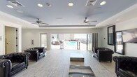 This rooftop space is thoughtfully designed to blend interior comfort with open-air living. The main indoor lounge area features light-toned tile flooring, recessed lighting, and dual ceiling fans that maintain a breezy atmosphere. Tufted leather club chairs are arranged around a low-profile coffee table, creating an inviting seating area. Large folding glass doors span the width of the room, opening fully to the outdoor pool deck and providing expansive city and sky views. Built-in floor-level water features add a subtle design element to the interior.

An adjacent lounge area offers a wall-mounted flat-screen television flanked by decorative mirrors, with additional leather seating and a tray ceiling detail accented in blue, adding a sophisticated touch. The wet bar area is outfitted with warm wood cabinetry, stainless steel countertops, a double sink, pendant lighting, and a pass-through mirror window connecting to the lounge.

Restrooms feature natural travertine tile flooring and striking black-and-white mosaic accent walls, lending a polished, boutique-style finish. The outdoor pool deck visible through the open doors completes this well-appointed rooftop retreat.
