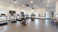 This fitness center in East Providence, Rhode Island, is a thoughtfully designed single-floor facility offering a comprehensive workout environment. The space is divided into two distinct zones, each catering to different exercise needs. The main cardio area features a row of treadmills and upright stationary bikes arranged across a broad, open floor finished in durable dark rubber flooring. Recessed lighting and dual ceiling fans maintain a bright, comfortable atmosphere, while multiple windows along the perimeter walls allow natural light to fill the space and provide views to the outdoors. Wall-mounted flat-screen televisions are positioned for convenient viewing during cardio sessions, and a large mirror along one wall enhances the sense of depth and allows for form monitoring.

A connected strength and functional training area houses a cable machine, a medicine ball rack stocked with a variety of weighted balls, resistance bands, a stability ball, and a rowing machine. This zone features protective rubber floor tiles and benefits from large windows that bring in ample natural light. The facility also includes a vending area near the entrance, restroom access, and a glass-paneled entry door, making it a fully self-contained and accessible workout destination.