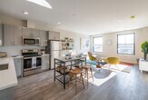 This well-designed single-floor residence offers a thoughtful layout that seamlessly blends modern aesthetics with functional living. The open-concept kitchen and living area serves as the heart of the unit, where a sleek kitchen outfitted with grey high-gloss cabinetry, white quartz countertops, stainless steel appliances — including a refrigerator, range, and microwave — flows naturally into the dining and living spaces. Light wood-tone flooring runs throughout, enhancing the bright, airy atmosphere. Large black-framed windows in the living area admit generous natural light and provide views of the surrounding neighborhood. The living room offers ample space for comfortable seating arrangements.

The unit includes two bedrooms, each carpeted and filled with natural light from oversized black-framed windows. The primary bedroom features direct access to a full bathroom and convenient proximity to the in-unit washer. A second full bathroom includes a walk-in shower with subway tile surround, a grey vanity with white countertop, and warm gold-toned fixtures. In-unit laundry is neatly tucked alongside the bathroom corridor. The overall design palette — neutral walls, modern finishes, and warm accent tones — creates a cohesive and inviting living environment throughout.