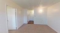 This single-floor, one-bedroom, one-bathroom unit offers a practical and well-organized layout designed to maximize functionality. The living area features neutral-toned carpet throughout, clean white walls, and a textured ceiling, creating a bright and open atmosphere. Wall-mounted lighting adds a subtle warmth to the space, while multiple electrical outlets are conveniently positioned throughout.

The bedroom is a comfortable, carpeted room equipped with a ceiling fan and a wide window fitted with horizontal blinds, allowing natural light to filter in along with views of the surrounding greenery and neighboring buildings. A wall-mounted HVAC unit provides independent climate control directly within the room.

The entryway and hallway transition seamlessly into the main living areas, with durable hardwood-style flooring providing a clean contrast to the carpeted rooms. The bathroom is thoughtfully designed with accessibility in mind, featuring stainless steel grab bars positioned near the bathtub and throughout the room, white tile surrounding the tub/shower combination, hardwood-style flooring, and a vanity with counter space.

Overall, this unit presents a clean, straightforward living space with practical finishes and accessible features, ideal for comfortable everyday living.
