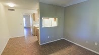 This single-story space offers a straightforward, functional layout with an open feel that connects the main living area to the kitchen. The living space is finished with durable tile-style flooring in a warm earth tone, complemented by neutral gray walls and white baseboards that create a clean, understated aesthetic. A defining architectural feature is the pass-through window cut into the dividing wall between the living area and the kitchen, promoting a sense of openness and allowing for easy communication between the two spaces. The kitchen is equipped with light wood cabinetry that extends to the upper walls, offering ample storage. A white refrigerator is visible through the pass-through, and the kitchen counters provide practical workspace. The textured ceiling adds a subtle design element consistent throughout the space. A hallway extending from the kitchen area leads toward additional rooms, with natural light visible from a window or door at the far end, suggesting good access to daylight. Overall, the space presents a practical and tidy interior with a cohesive color palette of warm neutrals and light wood tones.