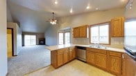 This single-floor space offers an open-concept layout that seamlessly connects the kitchen and living area, creating a spacious and cohesive environment. The kitchen is well-appointed with warm honey-toned oak cabinetry, a peninsula counter that doubles as a breakfast bar, a double-basin sink, and a dishwasher. Light tile flooring anchors the kitchen zone, while a transition to plush carpeting defines the living area. Multiple windows throughout the space invite generous natural light, offering views of the surrounding neighborhood. The living area features vaulted ceilings that lend an airy, expansive feel, further enhanced by a ceiling fan with integrated lighting. A fireplace serves as a focal point in the living room, adding a cozy, welcoming ambiance. Sliding glass doors open to a balcony, providing a direct connection to the outdoors. Recessed lighting throughout ensures consistent, well-distributed illumination. The neutral gray wall tones complement the warm wood finishes, creating a balanced and inviting atmosphere throughout the space.