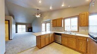 This single-floor space offers an open-concept layout that seamlessly connects the kitchen and living area, creating a spacious and cohesive environment. The kitchen is well-appointed with warm honey-toned oak cabinetry, a peninsula counter that doubles as a breakfast bar, a double-basin sink, and a dishwasher. Light tile flooring anchors the kitchen zone, while a transition to plush carpeting defines the living area. Multiple windows throughout the space invite generous natural light, offering views of the surrounding neighborhood. The living area features vaulted ceilings that lend an airy, expansive feel, further enhanced by a ceiling fan with integrated lighting. A fireplace serves as a focal point in the living room, adding a cozy, welcoming ambiance. Sliding glass doors open to a balcony, providing a direct connection to the outdoors. Recessed lighting throughout ensures consistent, well-distributed illumination. The neutral gray wall tones complement the warm wood finishes, creating a balanced and inviting atmosphere throughout the space.