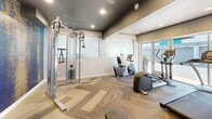 Located at 4341 Spring Street in La Mesa, California, this fitness center is a well-appointed exercise space designed to meet a variety of workout needs. The room features herringbone-patterned flooring that adds a stylish, contemporary touch, complemented by a dark ceiling with recessed lighting that creates a focused, energetic atmosphere.

The space is equipped with a comprehensive selection of fitness equipment, including multiple treadmills, an elliptical trainer, a recumbent bike, and a cable resistance machine — providing ample options for both cardio and strength training. A dedicated free weights area features a dumbbell rack with a selection of weights alongside a workout bench and a wall-mounted mirror, allowing users to monitor their form. A wall-mounted flat-screen television adds an entertainment element to the workout environment.

A striking blue and gold abstract accent wall serves as a vibrant focal point, lending artistic energy to the space. Large sliding glass doors and windows along one wall invite abundant natural light and provide views of an outdoor area, creating an open and airy feel despite the room's compact footprint. A whiteboard panel on one wall adds a functional element suitable for tracking workouts or fitness goals. The overall design balances practicality with modern aesthetics, making this fitness center an inviting and functional space for residents.