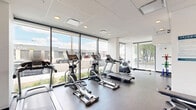 The Uptown Fitness Suite at 517 67 Ave SW in Calgary, AB is a thoughtfully designed, single-floor exercise facility offering a comprehensive range of equipment for residents and users alike. The space is defined by its clean, modern aesthetic, featuring a crisp white-and-grey colour palette, a drop-panel ceiling with recessed lighting, and durable rubber flooring throughout.

Floor-to-ceiling windows line much of the perimeter, flooding the space with natural light and offering open views of the surrounding streetscape and mature trees. This transparency creates an airy, motivating atmosphere that distinguishes the suite from a typical indoor gym.

The cardio zone is well-appointed, featuring multiple Spirit-brand treadmills, elliptical trainers, a stationary bike, and a rowing machine — accommodating a variety of aerobic workout preferences. A dedicated strength area houses a multi-functional cable machine, a weight plate storage tree with coloured bumper plates, and an adjustable workout bench, providing options for both resistance training and functional fitness.

The entry area offers additional workout stations and connects to the main exercise floor via a glass-paneled door. Informational signage, including facility rules and guidelines, is clearly posted on the walls. The overall layout is open and well-organized, making efficient use of the available floor area while maintaining a spacious, uncluttered feel suitable for a focused fitness experience.
