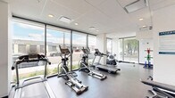 The Uptown Fitness Suite at 517 67 Ave SW in Calgary, AB is a thoughtfully designed, single-floor exercise facility offering a comprehensive range of equipment for residents and users alike. The space is defined by its clean, modern aesthetic, featuring a crisp white-and-grey colour palette, a drop-panel ceiling with recessed lighting, and durable rubber flooring throughout.

Floor-to-ceiling windows line much of the perimeter, flooding the space with natural light and offering open views of the surrounding streetscape and mature trees. This transparency creates an airy, motivating atmosphere that distinguishes the suite from a typical indoor gym.

The cardio zone is well-appointed, featuring multiple Spirit-brand treadmills, elliptical trainers, a stationary bike, and a rowing machine — accommodating a variety of aerobic workout preferences. A dedicated strength area houses a multi-functional cable machine, a weight plate storage tree with coloured bumper plates, and an adjustable workout bench, providing options for both resistance training and functional fitness.

The entry area offers additional workout stations and connects to the main exercise floor via a glass-paneled door. Informational signage, including facility rules and guidelines, is clearly posted on the walls. The overall layout is open and well-organized, making efficient use of the available floor area while maintaining a spacious, uncluttered feel suitable for a focused fitness experience.