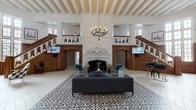 This remarkable single-floor space showcases an impressive blend of historic grandeur and contemporary functionality. The expansive main hall commands immediate attention with its soaring beamed ceiling, finished in crisp white with ornate decorative detailing, and illuminated by wrought-iron candelabra-style chandeliers that reinforce the space's classical character.

A striking black-and-white patterned encaustic tile floor anchors the central area, transitioning to a light herringbone wood floor that extends throughout the remainder of the hall. Symmetrical dual staircases with richly carved dark wood balustrades flank a grand white marble fireplace, creating a dramatic focal point. The mezzanine-level balustrade features distinctive decorative carved figures, adding a unique artistic element.

Tall multi-paned windows line the walls, flooding the interior with abundant natural light. Ornate dark wood columns with elaborately carved capitals punctuate the open floor plan, which accommodates distinct zones including a lounge seating area with tufted sofas and velvet armchairs near the fireplace, a billiards table, a glass-partitioned workspace, and casual dining furniture. The overall ambiance is one of distinguished, heritage-rich elegance with thoughtfully integrated modern conveniences.