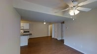 This single-floor residence offers a thoughtfully designed open-concept layout that seamlessly connects the kitchen and main living area. Rich, dark hardwood flooring runs throughout, adding warmth and a sense of continuity across the space. The living area is generously sized, offering ample room for comfortable furnishings, and is illuminated by a ceiling fan with an integrated light fixture that provides both style and functionality. A second overhead light further brightens the interior, keeping the space feeling open and well-lit. The kitchen is positioned along one side of the open floor plan and features white cabinetry, dark countertops, and standard appliances, including a range and overhead microwave. The contrast between the white cabinetry and dark counters gives the kitchen a clean, classic look. Neutral-toned walls provide a versatile backdrop throughout, complementing both the flooring and cabinetry. Multiple interior doors lead to additional rooms, suggesting a practical and well-organized floor plan. White baseboards and trim offer a crisp finishing detail throughout. Overall, the space presents a functional, inviting atmosphere suited for everyday living.