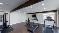 This single-floor home fitness space is thoughtfully designed to support an active lifestyle within a clean, well-appointed environment. The room's most striking architectural feature is its dark, exposed wood ceiling beams, which add warmth and character to the otherwise modern and neutral interior. Crisp white walls and bright white baseboards keep the space feeling fresh and open.

Underfoot, rich dark hardwood flooring runs throughout, providing a durable and attractive surface suited for a variety of workout activities. A generous array of recessed ceiling lights ensures the space is evenly and brightly illuminated at all times, creating an energizing atmosphere for exercise.

A wall-mounted flat-screen television is positioned prominently, allowing users to follow workout programs, stream entertainment, or monitor fitness metrics during training sessions. The room provides ample open floor space to accommodate multiple pieces of cardio equipment simultaneously, including treadmills and a stationary cycle, with room to spare for additional equipment or stretching areas. A window along one wall allows for natural light to filter in, adding to the overall sense of openness. The combination of purposeful design and practical amenities makes this a highly functional and inviting dedicated fitness room.