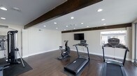 This single-floor home fitness space is thoughtfully designed to support an active lifestyle within a clean, well-appointed environment. The room's most striking architectural feature is its dark, exposed wood ceiling beams, which add warmth and character to the otherwise modern and neutral interior. Crisp white walls and bright white baseboards keep the space feeling fresh and open.

Underfoot, rich dark hardwood flooring runs throughout, providing a durable and attractive surface suited for a variety of workout activities. A generous array of recessed ceiling lights ensures the space is evenly and brightly illuminated at all times, creating an energizing atmosphere for exercise.

A wall-mounted flat-screen television is positioned prominently, allowing users to follow workout programs, stream entertainment, or monitor fitness metrics during training sessions. The room provides ample open floor space to accommodate multiple pieces of cardio equipment simultaneously, including treadmills and a stationary cycle, with room to spare for additional equipment or stretching areas. A window along one wall allows for natural light to filter in, adding to the overall sense of openness. The combination of purposeful design and practical amenities makes this a highly functional and inviting dedicated fitness room.