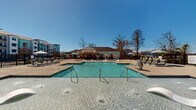 Located at 208 Windfall Way in Myrtle Beach, South Carolina, this outdoor amenity area is designed as a resort-style retreat for residents of the surrounding two-story community. At its centerpiece is a generously sized swimming pool with crystal-clear blue water, accessible via multiple stainless steel entry rails and featuring shallow wading ledges with in-pool loungers ideal for relaxing in the South Carolina sun.

The pool deck is finished in a warm tan concrete surface and is equipped with numerous chaise lounges and seating areas, including umbrella-shaded groupings and vibrant yellow accent chairs that add a lively coastal touch. A dedicated poolhouse structure anchors one end of the deck, clad in a distinctive aqua/turquoise siding with white trim and brick accents. This building houses restroom facilities, outdoor showers, and a water fountain, all accessible directly from the pool deck.

The entire area is enclosed by black wrought iron fencing, providing both security and an open visual aesthetic. Ceiling fans are installed under the covered porch of the poolhouse, enhancing comfort during warmer months. The surrounding landscape includes mature trees and manicured plantings, contributing to the welcoming, resort-inspired ambiance of this well-appointed outdoor space.