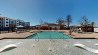 Located at 208 Windfall Way in Myrtle Beach, South Carolina, this outdoor amenity area is designed as a resort-style retreat for residents of the surrounding two-story community. At its centerpiece is a generously sized swimming pool with crystal-clear blue water, accessible via multiple stainless steel entry rails and featuring shallow wading ledges with in-pool loungers ideal for relaxing in the South Carolina sun.

The pool deck is finished in a warm tan concrete surface and is equipped with numerous chaise lounges and seating areas, including umbrella-shaded groupings and vibrant yellow accent chairs that add a lively coastal touch. A dedicated poolhouse structure anchors one end of the deck, clad in a distinctive aqua/turquoise siding with white trim and brick accents. This building houses restroom facilities, outdoor showers, and a water fountain, all accessible directly from the pool deck.

The entire area is enclosed by black wrought iron fencing, providing both security and an open visual aesthetic. Ceiling fans are installed under the covered porch of the poolhouse, enhancing comfort during warmer months. The surrounding landscape includes mature trees and manicured plantings, contributing to the welcoming, resort-inspired ambiance of this well-appointed outdoor space.