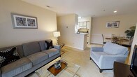 This single-floor residence offers a well-designed open-concept layout that seamlessly connects the living area to the kitchen, maximizing the sense of space and ease of movement throughout. The living room is carpeted in a warm neutral tone and provides ample room for seating, as evidenced by the plush sofa and accent chair arrangement centered around a glass-top coffee table. Soft, beige walls are carried consistently throughout the space, contributing to a calm and cohesive ambiance. A framed piece of wall art adds a decorative touch to the living area, while a table lamp provides warm accent lighting. The kitchen is positioned at the rear of the living space and is accessible via a peninsula-style breakfast bar, which creates a natural division between the two areas while maintaining the open feel. The kitchen features white cabinetry, overhead lighting, and essential appliances, offering a functional cooking environment. The pass-through design of the kitchen enhances the social and visual connection between cooking and living areas. Overall, the space is characterized by its neutral color palette, soft carpeting, and practical layout, making it a comfortable and inviting single-floor home.