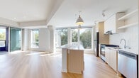 This single-floor residence showcases a contemporary design defined by clean lines, a neutral palette, and an abundance of natural light. The open-concept living and kitchen area serves as the heart of the unit, where light hardwood flooring extends seamlessly throughout, complementing the crisp white walls and ceilings. Large windows line the living area, framing views of surrounding greenery and flooding the space with sunlight.

The kitchen is thoughtfully designed with flat-front cabinetry in a warm wood tone paired with white uppers, a textured subway tile backsplash, and stainless steel appliances including a gas range, refrigerator, and dishwasher. A central island with a white countertop provides additional workspace and anchors the room. A modern pendant light fixture adds a stylish focal point above the island.

The bedrooms are well-proportioned, featuring the same warm hardwood flooring, neutral walls, and natural light from windows. Two bathrooms serve the unit: one features a floating white vanity with a large mirror, while the other includes a shower with tiled surround. Both maintain the clean, minimalist aesthetic consistent throughout the space.