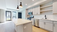 This single-floor space showcases a clean, modern design with thoughtful finishes throughout. The heart of the unit is an open-concept kitchen, anchored by a central island with a crisp white quartz countertop and built-in outlets, ideal for both meal prep and casual dining. A pair of striking black geometric pendant lights hang above, adding a stylish contrast to the light-toned cabinetry featuring matte black hardware. The kitchen is fully equipped with stainless steel appliances, including a gas range, microwave, dishwasher, and refrigerator. A vertical subway tile backsplash, open shelving, and a matte black faucet at the undermount sink complete the contemporary aesthetic. Light wood-tone flooring runs throughout the kitchen and living areas, enhancing the bright and airy atmosphere created by recessed lighting and large windows. The bathroom continues the modern theme with a floating vanity, white countertop, large mirror, and matte black fixtures, all set against clean white walls and warm wood-tone flooring. The bedroom offers a quiet retreat with soft carpeting, a ceiling fan with integrated lighting, and a window that frames a view of surrounding greenery, filling the room with natural light.