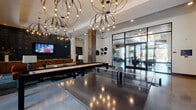 Located along Belt Line Road in Addison, Texas, this single-floor amenity space is designed with a contemporary aesthetic that blends industrial and upscale elements seamlessly. Polished concrete floors extend throughout, reflecting light from dramatic oversized globe chandeliers that serve as striking focal points in the main lounge and dining areas.

The game room is a standout feature, equipped with a ping-pong table, shuffleboard, a mounted flat-screen television, and an arcade game console, all set beneath sculptural lighting. Adjacent areas transition smoothly into comfortable lounge seating, where floor-to-ceiling black-framed glass walls and doors offer unobstructed views of a resort-style pool courtyard, flooding the interior with natural light.

The kitchen area is finished with granite and stone countertops, rich wood cabinetry, and a bold geometric tiled backsplash, complemented by a stainless steel refrigerator and large windows overlooking the outdoor amenity deck.

A glass-enclosed conference or co-working room provides a more focused environment, while a formal private dining area features a long dark table, leather-upholstered seating, an ethanol fireplace set into a textured decorative wall, and additional globe lighting overhead. Reclaimed wood accent columns add warmth and texture throughout the space.