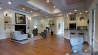 This well-appointed single-floor space offers an open, airy layout that immediately impresses with its sense of scale and refined design. The main living area features rich hardwood flooring that extends throughout, complemented by a striking coffered ceiling with recessed lighting that adds depth and a polished architectural character to the room. The space is bathed in a warm, welcoming ambiance, enhanced by carefully placed accent lighting and an abundance of natural light filtering in through multiple windows and a glass-paneled door visible in the background.

The living area is thoughtfully arranged with comfortable seating, including tufted armchairs and a sofa, centered around a coffee table atop a patterned area rug — creating an inviting lounge setting. A built-in shelving unit and fireplace anchor one wall, providing both functionality and a classic focal point.

A graceful arched passageway leads to additional areas of the space, including what appears to be a dedicated workspace or office room. Adjacent to the main living area, a raised counter with bar-height seating and a sleek, dark countertop defines a casual dining or refreshment zone. A tall potted plant adds a vibrant natural element, tying the interior together with an organic touch throughout this sophisticated and versatile space.