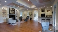 This well-appointed single-floor space offers an open, airy layout that immediately impresses with its sense of scale and refined design. The main living area features rich hardwood flooring that extends throughout, complemented by a striking coffered ceiling with recessed lighting that adds depth and a polished architectural character to the room. The space is bathed in a warm, welcoming ambiance, enhanced by carefully placed accent lighting and an abundance of natural light filtering in through multiple windows and a glass-paneled door visible in the background.

The living area is thoughtfully arranged with comfortable seating, including tufted armchairs and a sofa, centered around a coffee table atop a patterned area rug — creating an inviting lounge setting. A built-in shelving unit and fireplace anchor one wall, providing both functionality and a classic focal point.

A graceful arched passageway leads to additional areas of the space, including what appears to be a dedicated workspace or office room. Adjacent to the main living area, a raised counter with bar-height seating and a sleek, dark countertop defines a casual dining or refreshment zone. A tall potted plant adds a vibrant natural element, tying the interior together with an organic touch throughout this sophisticated and versatile space.