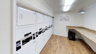 This shared laundry facility is designed with functionality and comfort in mind. The room features an extensive row of white commercial-grade washing machines and dryers lining one wall, including both stacked and front-loading units with digital controls, providing ample capacity for multiple users. A granite-topped folding counter offers a convenient surface for handling laundry.

The flooring throughout the space consists of light-toned hardwood-style planks, lending a clean and modern aesthetic. Crown molding trims the ceiling, adding a refined architectural detail that elevates the utilitarian nature of the room. Bright fluorescent panel lighting illuminates the space evenly, ensuring excellent visibility.

A slatted wooden bench positioned against the wall offers a comfortable place to wait. Informational signage is posted near the machines, providing usage instructions and guidelines. A doorway at the far end of the room connects to an adjacent hallway, suggesting convenient access within the broader building. The overall ambiance is clean, bright, and well-maintained, reflecting a practical yet thoughtfully designed communal space.
