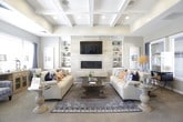 This single-story community space offers a polished and welcoming environment designed for both comfort and functionality. The entrance features a glass-paneled double-door entry with a vaulted ceiling, large entry mat, and tile flooring that sets an impressive first impression.

The central foyer is a standout architectural highlight, boasting soaring vaulted ceilings with white exposed beams and a striking chandelier. Decorative gold mirror accents adorn the walls, while light tile flooring and an area rug anchor the open layout. Doorways lead seamlessly to multiple adjoining spaces, including a private office with built-in shelving, a ceiling fan, and a dedicated desk area, as well as a cozy sitting nook bathed in natural light.

The lounge area is generously proportioned and thoughtfully designed, featuring a coffered ceiling, large windows dressed with navy drapes and plantation shutters, and an abundance of natural light. Built-in shelving flanks a sleek tiled fireplace with a wall-mounted television above. Comfortable seating, layered area rugs, a glass-top coffee table, and eclectic decorative accents complete the inviting atmosphere. The warm, neutral color palette with bold accent tones throughout creates a cohesive and sophisticated ambiance across the entire space.