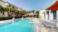 This outdoor amenity space centers around a expansive, crystal-clear swimming pool with a shimmering turquoise finish that evokes a resort-like ambiance. The pool features a clean, modern design with smooth tiled surrounds and decorative mosaic-accented elements that add a touch of elegance. A generous pool deck lines one side of the water, furnished with a row of cushioned lounge chairs ideal for sunbathing and relaxation.

Tall, mature palm trees are thoughtfully positioned throughout the area, providing natural shade and lending a distinctly tropical character to the setting. A covered pergola structure anchors the far end of the space, offering a shaded gathering area outfitted with additional seating, creating an inviting spot for socializing. Accent globe lighting and orange-cushioned lounge furniture introduce pops of color against the predominantly white and neutral palette of the surrounding building facades.

The surrounding low-rise architecture features light-toned stucco exteriors with large windows and clean lines, reinforcing a contemporary coastal aesthetic. Under open skies, the entire area benefits from abundant natural light, contributing to a bright, airy, and welcoming outdoor environment.