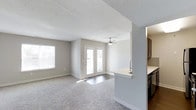 This single-floor residence offers a bright, open layout designed for comfortable everyday living. The main living area is generously proportioned, featuring plush wall-to-wall carpeting underfoot and a textured ceiling overhead. A ceiling fan centered in the room provides year-round comfort and airflow. Natural light fills the space through multiple windows fitted with horizontal blinds, as well as a set of French doors that open to the outside, adding an airy, welcoming quality to the room.

The kitchen is positioned adjacent to the living area in an open-concept arrangement, separated by a partial wall with a peninsula countertop that doubles as a casual dining or prep surface. Dark wood cabinetry provides ample storage, and a sink is conveniently integrated into the peninsula. The kitchen's proximity to the living space makes it well-suited for entertaining or day-to-day interaction between rooms.

A hallway connects the living space to the remaining areas of the home, featuring a sliding panel door—a practical and space-conscious design choice. An electrical panel is accessible along the hallway wall. The overall palette of neutral tones throughout creates a clean, cohesive, and move-in-ready environment.