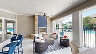 This generously sized single-floor covered outdoor lounge serves as a premier communal amenity space, seamlessly blending indoor comfort with open-air living. The centerpiece of the space is a dramatic floor-to-ceiling fireplace clad in stacked stone with a striking blue mosaic tile accent, flanked by a raised hearth and surrounded by wicker lounge chairs and a patterned area rug — creating an inviting focal point for gathering.

The layout opens broadly toward a pool area through wide, column-framed openings fitted with wrought iron railings, allowing unobstructed views of the aquatic amenities and lush landscaping beyond. Multiple ceiling fans are mounted throughout, ensuring comfortable airflow across the entire covered area.

Adjacent to the lounge seating, a curved granite-topped bar counter stretches along one side, lined with navy blue rope bar stools and illuminated by pendant lantern lights hanging overhead — a well-appointed spot for casual dining or socializing. Further into the space, additional seating arrangements include a sofa grouping on a geometric rug alongside high-top bar tables with coordinating navy chairs.

The exterior walls feature classic horizontal shiplap siding in a neutral taupe finish, and the space is accessible via a glass-paneled entry door. The overall ambiance is relaxed yet refined, combining coastal-inspired design with functional, resort-style amenities.