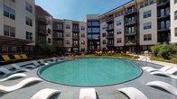 This contemporary multi-story residential community is designed around an impressive central courtyard that anchors the amenity experience. At its heart sits a large, circular resort-style swimming pool surrounded by a generous pool deck furnished with sleek white lounge chairs and vibrant yellow seating areas, creating an inviting outdoor retreat. The building's facade wraps around the courtyard in a curved configuration, with multiple floors of private balconies overlooking the pool area. The exterior architecture blends neutral tones with dark accent elements and expansive windows, lending a modern, upscale aesthetic throughout.

The fitness amenities are equally comprehensive. A spacious main gym features rows of treadmills and strength-training equipment set on patterned carpet flooring beneath bright, recessed lighting, with floor-to-ceiling glass walls providing an open, airy feel. Adjacent to the main floor is a dedicated studio outfitted with cycling bikes, a full wall of mirrors, colorful free weights, stability balls, and resistance bands for a complete functional workout experience. A large mounted display screen streams on-demand fitness content, further enhancing the training environment. Wood-look flooring, neutral gray walls, and a ceiling fan complete the studio's clean, focused design.