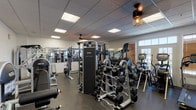 Located at 3101 Old 63 S in Columbia, Missouri, this fitness center offers a comprehensive, well-organized workout facility designed to support a full range of exercise routines. The single-floor space is thoughtfully laid out across multiple interconnected zones, each dedicated to different types of training.

The strength training area is stocked with an impressive array of equipment, including Nautilus Instinct selectorized machines, a functional trainer, a chin/dip assist station, plate-loaded machines, and a well-stocked dumbbell rack with a variety of weights. Floor-to-ceiling mirrors line the walls, enhancing the sense of space and allowing users to monitor their form.

The cardio section features treadmills, elliptical trainers, and stair climbers positioned near windows with wood-slat blinds, providing natural light and an airy atmosphere. Wall-mounted televisions are available to keep members entertained during cardio sessions.

The entryway includes a tiled foyer with a physician-style standing scale, offering a clean and functional transition into the workout area. A water cooler station is also available for member convenience.

Throughout the facility, durable rubber flooring provides safety and noise reduction, while a drop ceiling with recessed panel lighting and stylish cage pendant fixtures ensures consistent, well-distributed illumination. The overall ambiance is clean, bright, and professional.