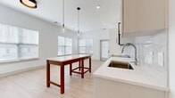 Located at 1200 Marquis Pkwy in Williamsburg, VA, this well-appointed 2-bedroom, 2-bathroom residence offers a thoughtfully designed single-floor layout. The open-concept kitchen and living area serves as the heart of the home, featuring crisp white cabinetry extending to the ceiling, stainless steel appliances including a refrigerator, range, microwave, and dishwasher, and a white subway tile backsplash. A large kitchen peninsula with a stainless steel sink provides additional workspace, while stylish pendant lighting adds a modern touch overhead. The living area is bathed in natural light from multiple windows and features light-toned hardwood-style flooring and elevated ceilings that enhance the sense of openness. Both bedrooms are generously sized, carpeted for comfort, and receive ample natural light through wide windows. The primary bedroom includes access to a dedicated closet with built-in wire shelving. The two bathrooms are finished in a clean, contemporary style with white vanities, black hardware accents, light wood-style flooring, and framed mirrors. One bathroom includes a full tub-shower combo with white tile surround. Hallways connect the spaces efficiently, and the overall palette of white walls and light finishes creates a bright, cohesive ambiance throughout.