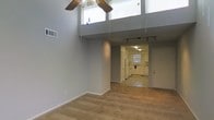 This single-floor space makes a strong first impression with its dramatic double-height ceilings in the main living area, which create an expansive, airy atmosphere. Large upper-level windows allow natural light to filter down into the space, enhancing the open feel. A ceiling fan with a light fixture is centrally positioned, adding both function and a decorative element overhead.

The living area features neutral-toned carpet flooring and smooth gray walls, providing a clean, versatile backdrop. Track lighting runs along the ceiling toward the back of the space, offering adjustable illumination throughout. The open layout flows naturally from the living area into the kitchen, which transitions to tile flooring.

The kitchen is equipped with white cabinetry, white appliances including a range, and a sink positioned beneath a window that brings in additional natural light. The cabinetry extends along the walls, providing ample storage. A second door is visible near the kitchen, likely serving as an exterior entry point.

Overall, the space balances functional design with an open, light-filled ambiance, highlighted by the striking vertical volume of the main living area.