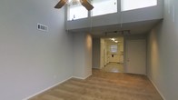 This single-floor space makes a strong first impression with its dramatic double-height ceilings in the main living area, which create an expansive, airy atmosphere. Large upper-level windows allow natural light to filter down into the space, enhancing the open feel. A ceiling fan with a light fixture is centrally positioned, adding both function and a decorative element overhead.

The living area features neutral-toned carpet flooring and smooth gray walls, providing a clean, versatile backdrop. Track lighting runs along the ceiling toward the back of the space, offering adjustable illumination throughout. The open layout flows naturally from the living area into the kitchen, which transitions to tile flooring.

The kitchen is equipped with white cabinetry, white appliances including a range, and a sink positioned beneath a window that brings in additional natural light. The cabinetry extends along the walls, providing ample storage. A second door is visible near the kitchen, likely serving as an exterior entry point.

Overall, the space balances functional design with an open, light-filled ambiance, highlighted by the striking vertical volume of the main living area.