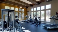 This fitness center is designed to provide an energizing and comfortable workout environment. The space is defined by its impressive ceiling height, which is accentuated by decorative wood beam detailing and recessed lighting, creating a structured yet open atmosphere. A ceiling fan adds to the room's ventilation and comfort.

Natural light is a standout feature, with large multi-pane windows lining two walls and extending nearly floor to ceiling. These windows offer views of mature trees and the surrounding outdoor landscape, contributing to a bright and motivating ambiance. The warm golden-yellow wall tones complement the dark wood beam accents, lending the space a polished, welcoming aesthetic.

The fitness center is equipped with a variety of cardio machines, including treadmills, ellipticals, a stationary bike, and rowing equipment, all arranged to maximize space and usability. Strength training equipment is also present along the perimeter. The dark rubber flooring provides durability and safety underfoot. Overall, the space balances functionality with thoughtful design, offering a well-lit, spacious environment suited for a full range of fitness activities.