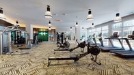 This well-appointed fitness center is designed to support a comprehensive workout experience across a generously sized, single-floor layout. The main exercise area is flooded with natural light through expansive floor-to-ceiling windows that line the perimeter, offering views of an outdoor courtyard and surrounding building. Stylish pendant lights with an industrial aesthetic hang from white ceilings, complementing the bold geometric-patterned carpet that runs throughout the space.

The cardio zone is equipped with multiple treadmills, ellipticals, and Concept2 RowERG rowing machines, providing ample options for endurance training. A separate, glass-enclosed weight room features a striking green accent wall adorned with a neon heartbeat graphic, along with an array of strength machines, free weights, dumbbells, and a power rack — all set beneath a warm wood-paneled ceiling with recessed lighting that creates a focused, high-performance atmosphere.

Large mirrors are strategically placed throughout to enhance the sense of space and aid in form monitoring. A glassed entry vestibule with a warm wood ceiling ceiling serves as a transition point, with direct access to the outdoor terrace. The overall ambiance is modern and motivating, combining upscale design elements with functional, professional-grade equipment to create an inviting environment for all fitness levels.