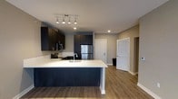 This single-floor space showcases a contemporary open-concept layout centered around a well-appointed kitchen. Dark espresso-toned cabinetry lines the walls, providing ample storage and a striking contrast against the light, neutral-toned walls. The kitchen is equipped with stainless steel appliances, including a refrigerator and range with microwave above, all complemented by crisp white countertops that extend across the peninsula. The peninsula, finished with a dark base panel, offers additional workspace and casual seating potential while serving as a natural divider between the kitchen and the adjoining living area. A track lighting fixture overhead provides focused illumination throughout the kitchen space, supplemented by recessed lighting for an even, well-lit environment. The flooring throughout is a warm-toned wood-look laminate, adding continuity and a welcoming feel across the open space. Two doorways visible from the main area lead to additional rooms, suggesting a functional and thoughtfully arranged floor plan. Overall, the space balances modern aesthetics with practical design elements.
