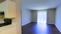 Located at 550 Jackson St. in Daphne, Alabama, this single-floor space offers a clean, straightforward layout designed for comfortable everyday living. The living area is a standout feature, showcasing rich, dark hardwood flooring that extends throughout the open space, providing a warm and polished aesthetic. A full-height sliding glass door dominates one wall of the living area, allowing an abundance of natural light to pour in while offering direct access to an outdoor area beyond. Vertical blinds accompany the sliding door, providing flexible light and privacy control. The textured ceiling adds a classic architectural detail that complements the neutral wall tones, creating a cohesive and understated interior palette. The transition from the adjacent kitchen area — visible with white cabinetry — into the living space reflects an open and functional flow. Clean white baseboards frame the perimeter of the room, adding a crisp finishing touch. The overall ambiance of the space is bright, airy, and move-in ready, making it a practical and inviting environment in the heart of Daphne, AL.